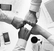 Four hands joining together in unity over paperwork and mobile devices on a white surface
