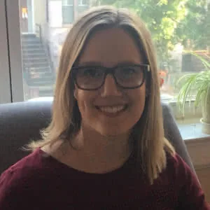Woman with long brown hair and dark-framed glasses smiling at camera, wearing maroon sweater indoors