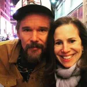 Two people smiling at camera on a city street at night with bright lights and buildings in background