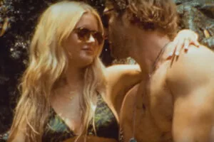 Woman wearing sunglasses and bikini top facing shirtless man outdoors near rocky surface