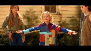 Three children standing outdoors in front of a wooden house, with the center child wearing a colorful patchwork jacket with arms outstretched.