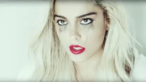 Woman with platinum blonde hair, bold pink lipstick, and dark eye makeup with tear-like markings below eyes