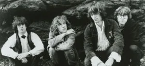 Four men with 1980s-style hair posed against a stone wall in black and white photograph