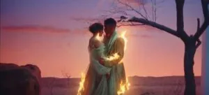 Two figures in light-colored clothing embrace under a bare tree against a dramatic sunset sky with golden light effects.