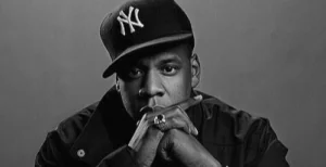 Man wearing black Yankees cap and jacket, hand raised to face in contemplative pose, black and white portrait