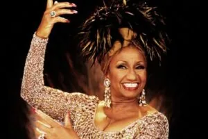 Woman in gold sequined dress with black feathered headpiece, pearl earrings, and diamond ring, smiling with raised hand against dark background
