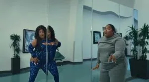 Two women in a modern office hallway with glass panels and potted plants, one wearing a blue sequined outfit touching a mirror, the other in gray laughing
