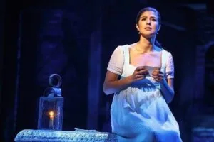 Woman in white dress on stage holding candle, looking upward with blue lighting