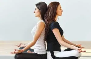 Two women sitting back-to-back on a wooden floor in meditation pose with hands resting on knees
