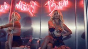 Woman in orange top with blonde hair sitting against mirrored wall with red neon signs above