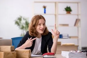 Woman in black blazer at desk gesturing excitedly with cardboard boxes, office shelving background