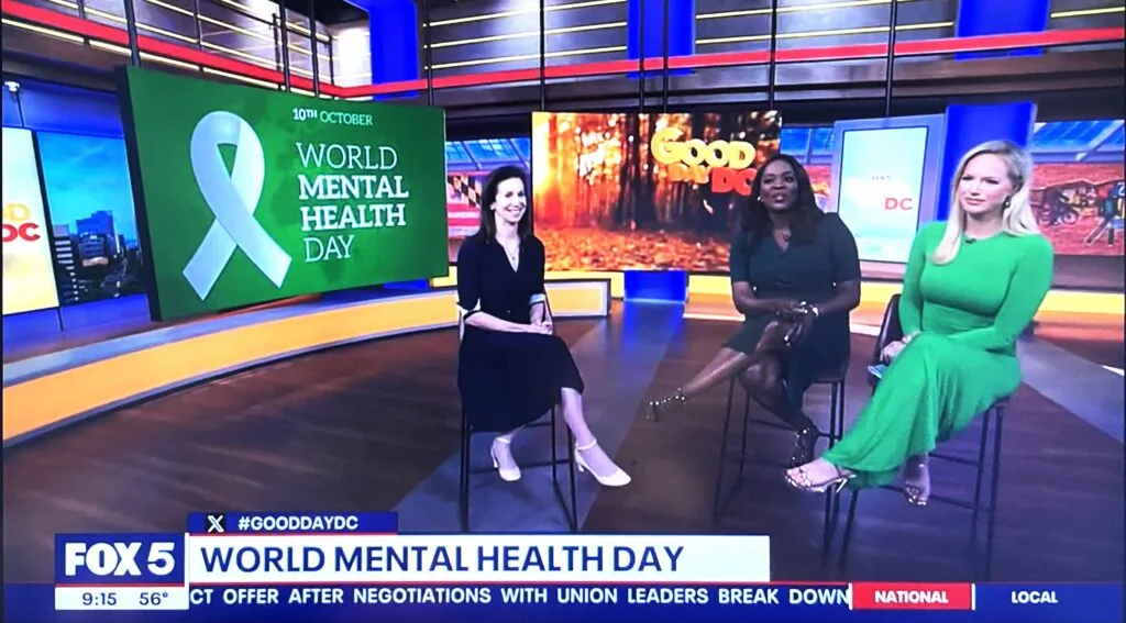 Three women seated on a television studio set with a green banner displaying a white ribbon and text reading '10th October World Mental Health Day'